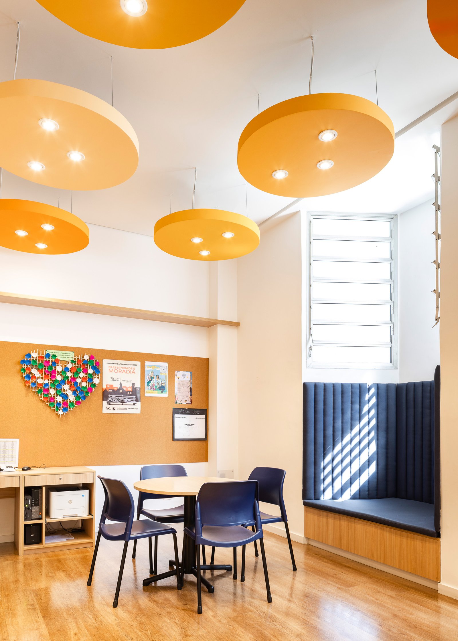 Sala da pastoral com pé-direito alto, luminárias em formato de nuvens e nicho lateral convertido em banco estofado azul