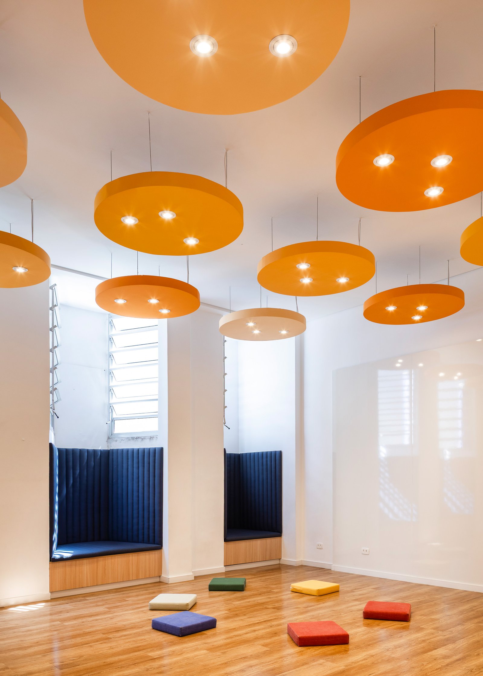 Ambiente coletivo com luminárias suspensas laranja, nichos estofados azuis e almofadas dispostas no piso de madeira