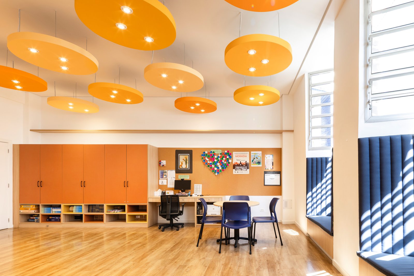 Sala da pastoral com grande armário laranja de organização, nichos abertos com jogos e livros e área de trabalho do coordenador