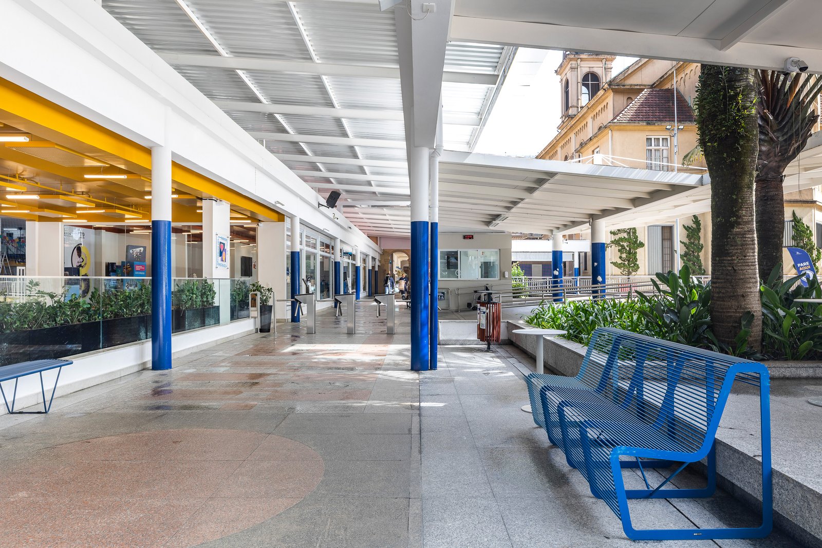 Passeio coberto do colégio com bancos azuis e a torre histórica ao fundo
