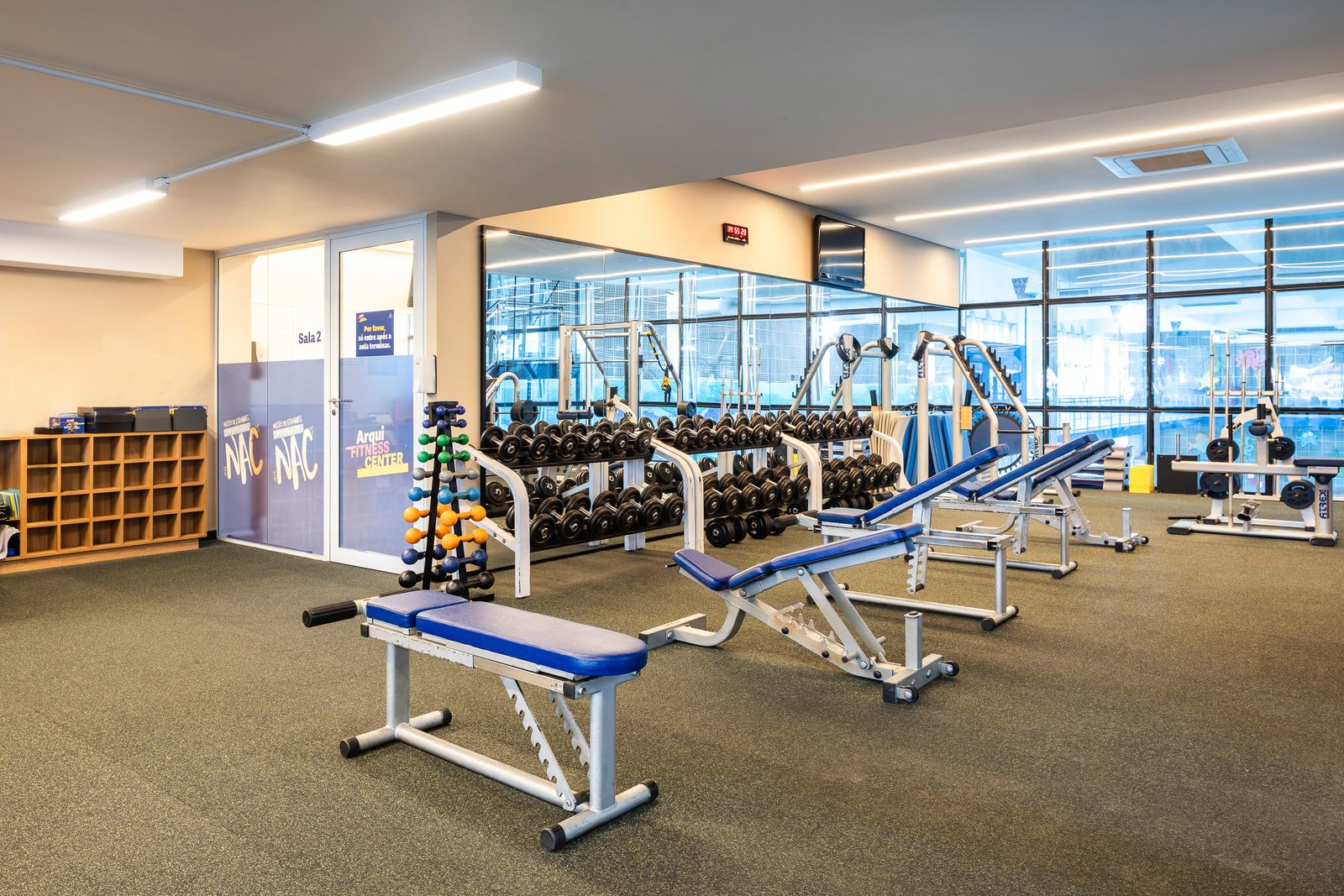 Sala de musculação entregue do Arqui Fitness Center, com halteres, bancos azuis e a porta de acesso à Sala 2