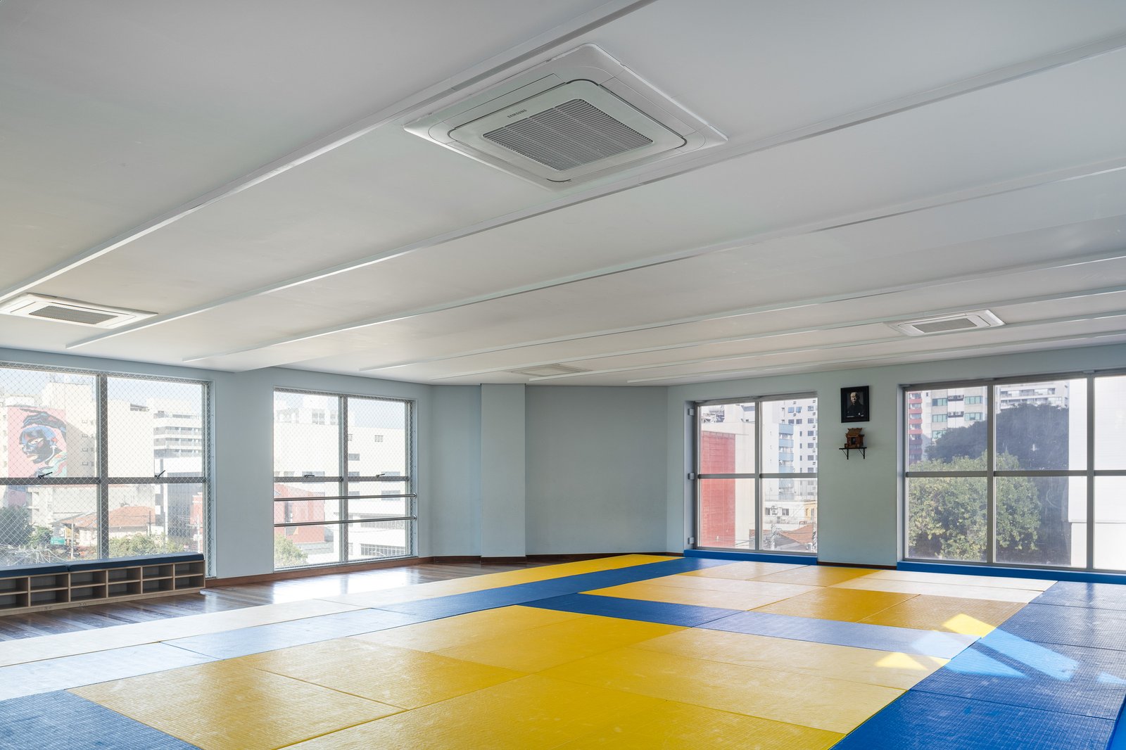 Sala de judô do NAC entregue, com tatames profissionais em azul e amarelo, vista para a cidade e nichos para calçados