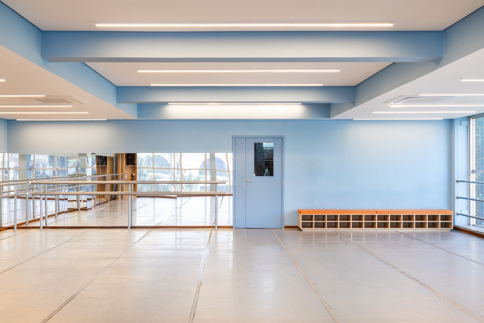 Sala de dança em azul claro, com barra, espelhos, bancos e nichos de organização