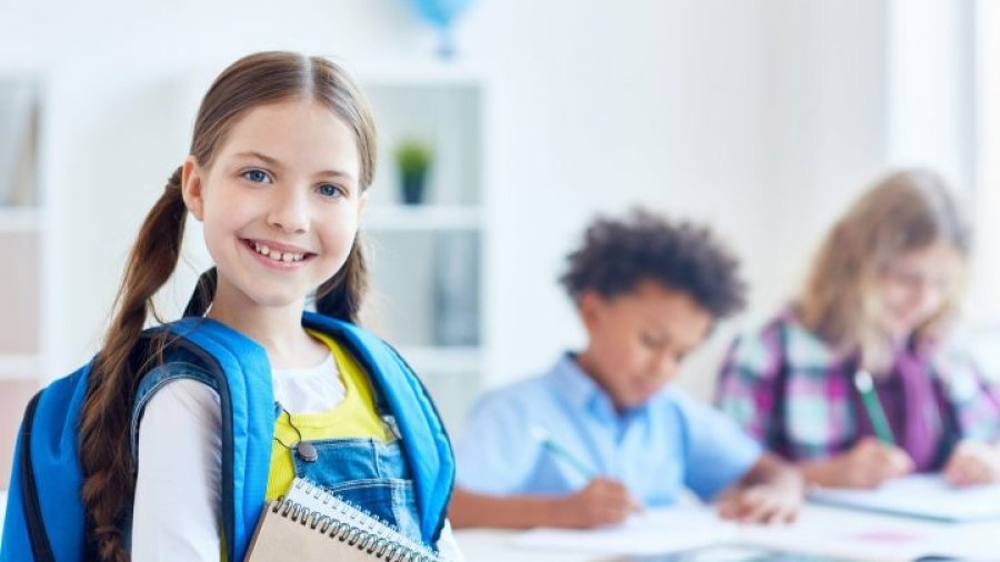 Smiling little scholar with rucksack and notepads on background of schoolboys