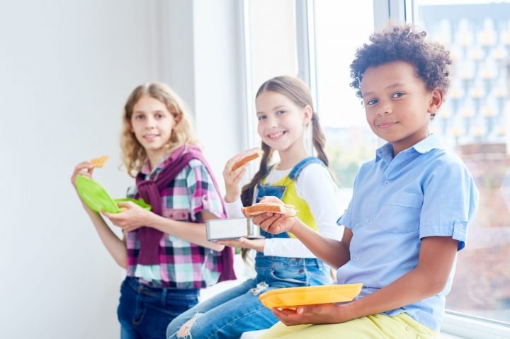 Adorable school learners with containers eating sandwiches on windowsill